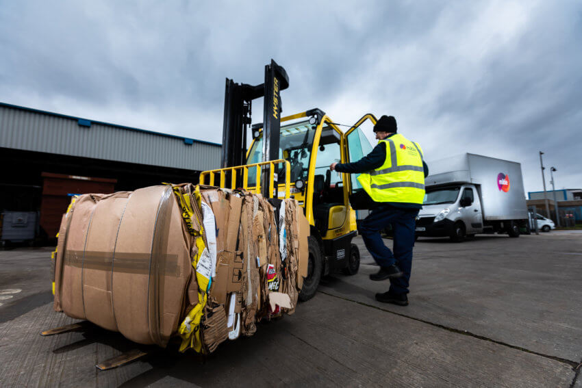 A man in a Mitie hi-vis vest climbing into a yellow forklift loaded with cardboard waste