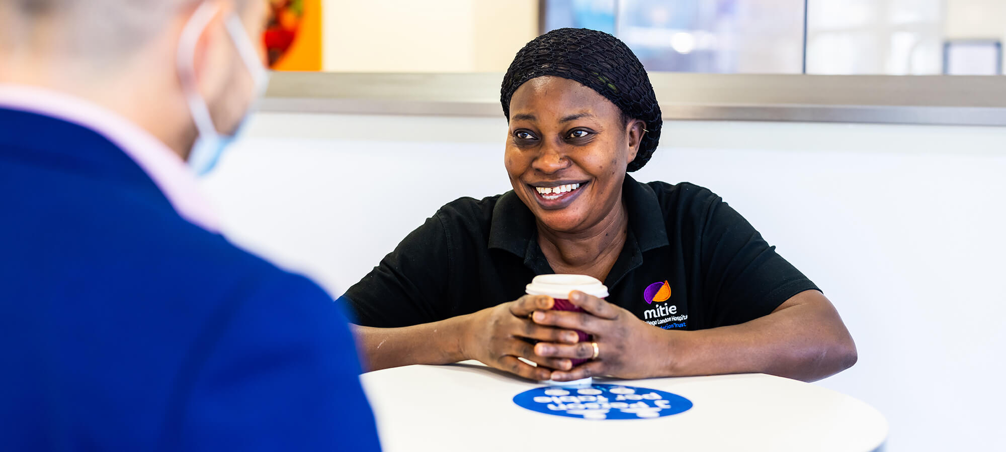 Smiling Mitie healthcare employee in a company branded black polo shirt, holding a cup of coffee while leaning on a white table