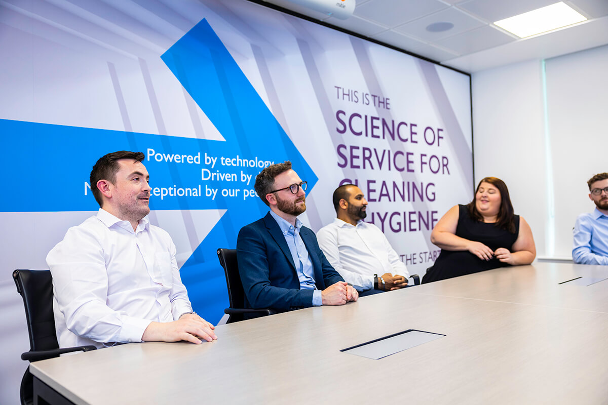 A group of people sitting by a table in a Mitie meeting room with 'This is the Science of Service for Cleaning & Hygiene' on the wall behind them