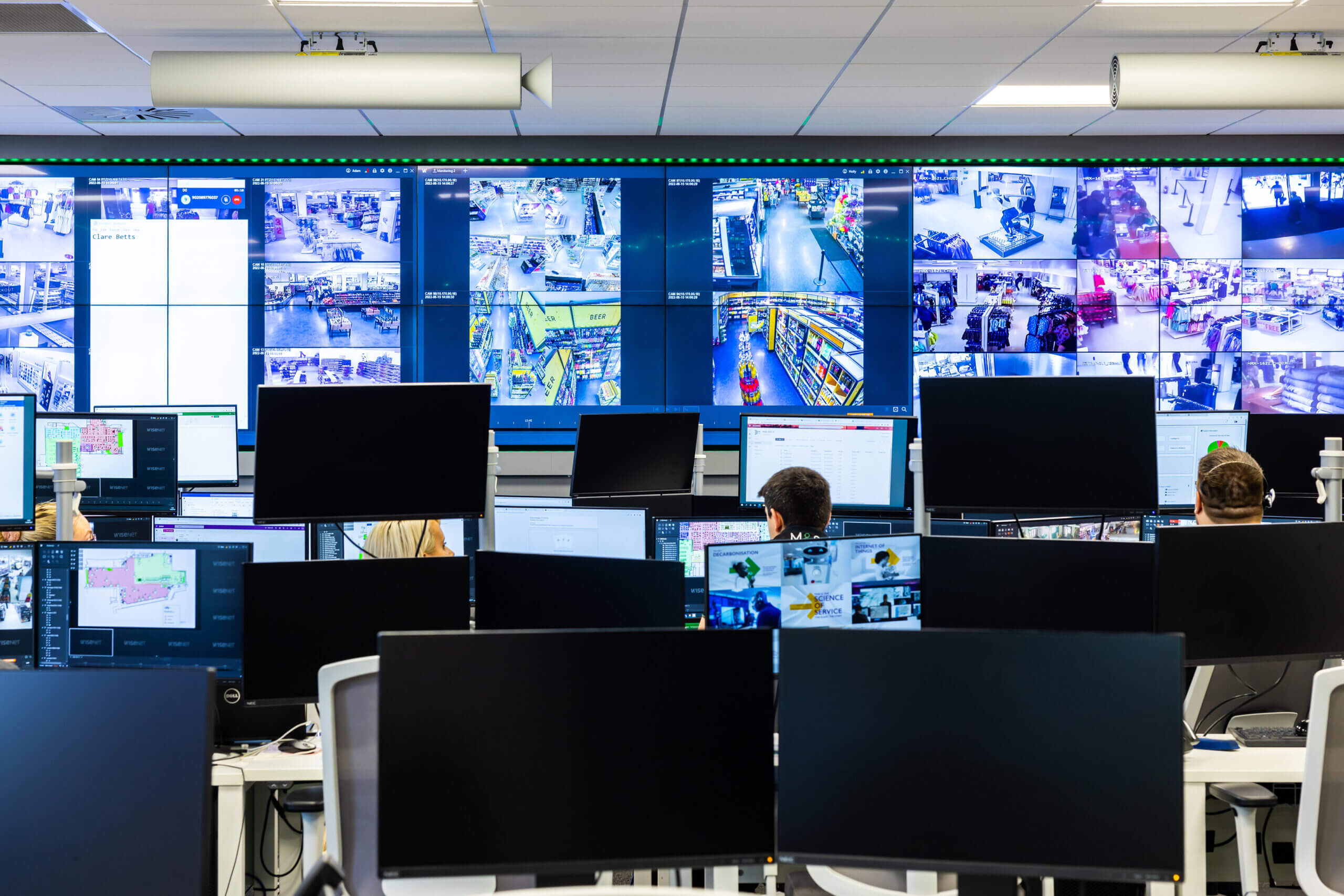 Various people working at desks with a wall of computer screens in front of them, showing a range of CCTV imagery